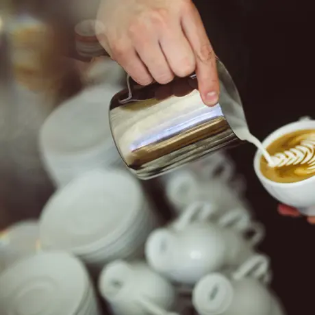 Barista nalewa mleko do filiżanki z latte artem, w tle stosy porcelany