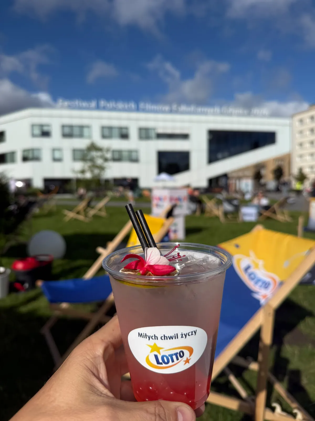 Różowa lemoniada bubble z kulkami na dnie, kwiatem i suszonym cytrusem w kubku z logo klienta, w tle leżaki w strefie festiwalowej