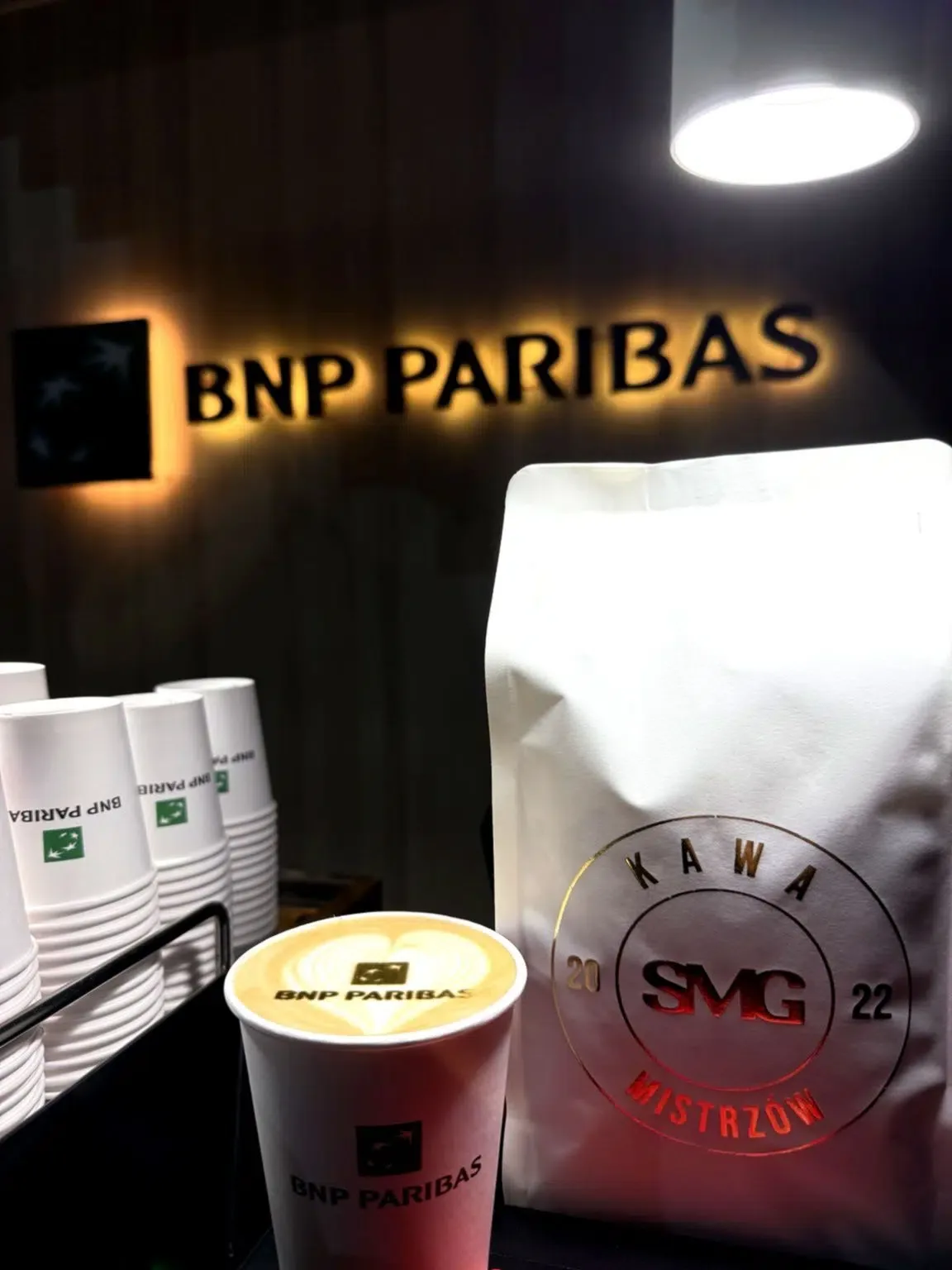 Kubek z logo BNP Paribas na piance, obok paczka ziaren i kubki na stoisku