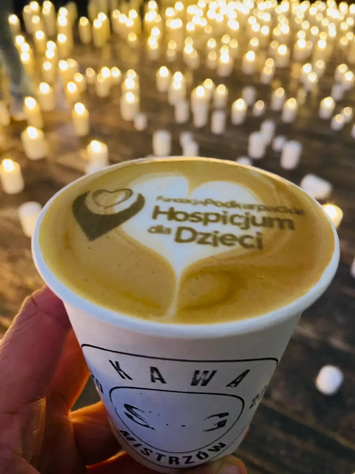 Kubek kawy z logo Hospicjum dla Dzieci na piance, w tle morze zniczy