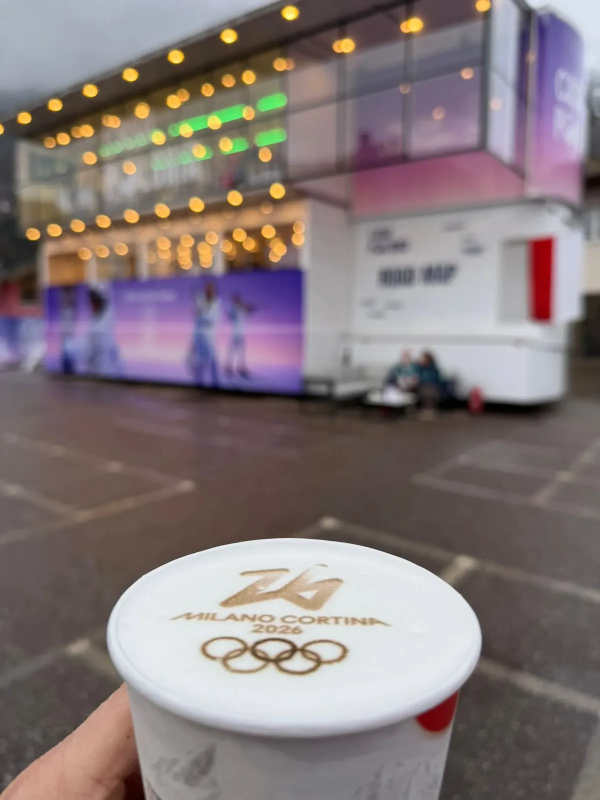 Kubek z logo igrzysk Milano Cortina 2026 na piance, w tle pawilon