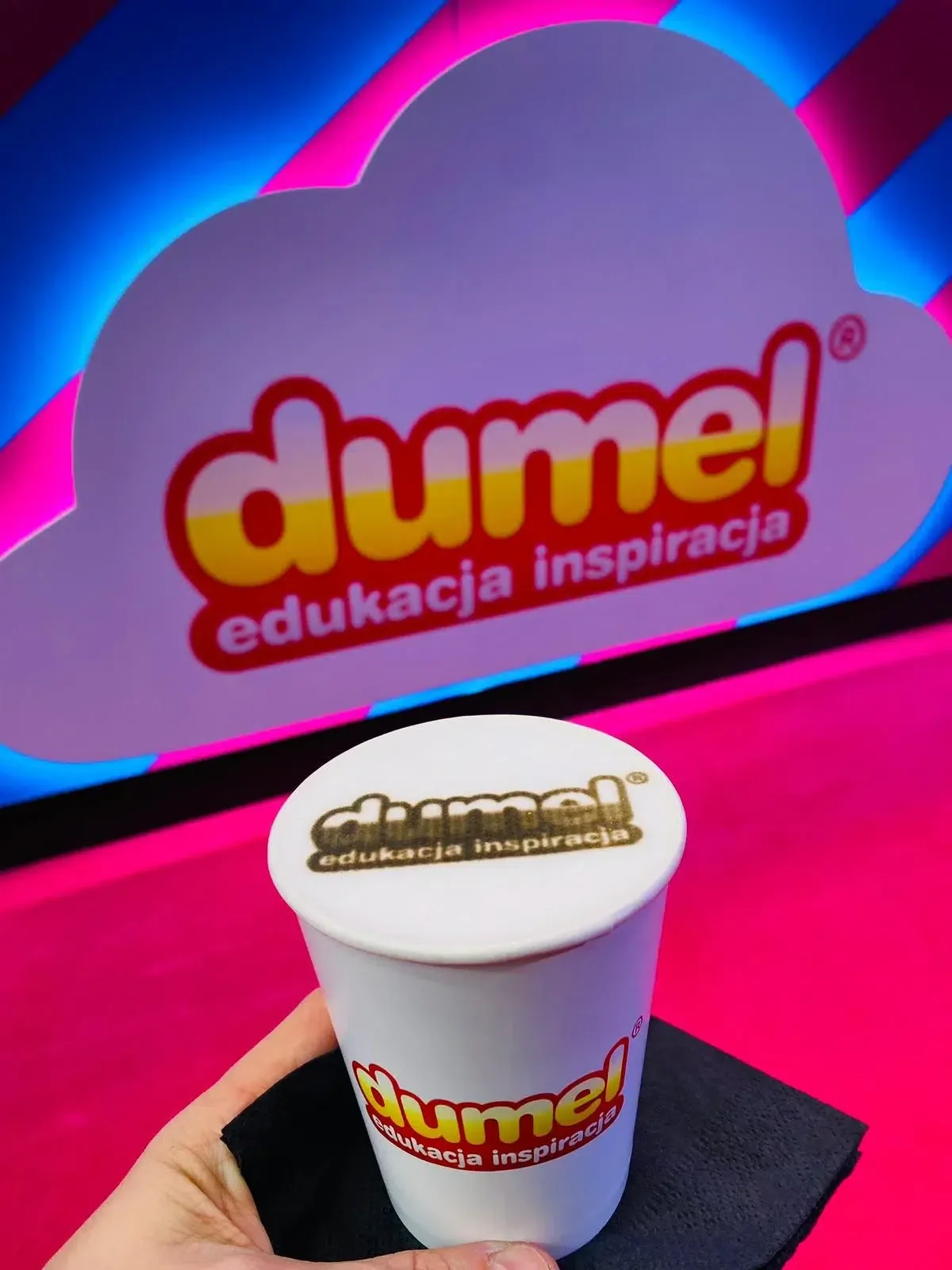Kubek z wydrukowanym logo marki Dumel na piance, na tle firmowego banera