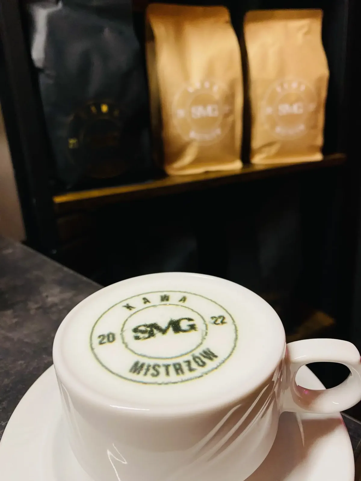 Filiżanka z logo SMG na piance, na spodku, w tle paczki ziaren