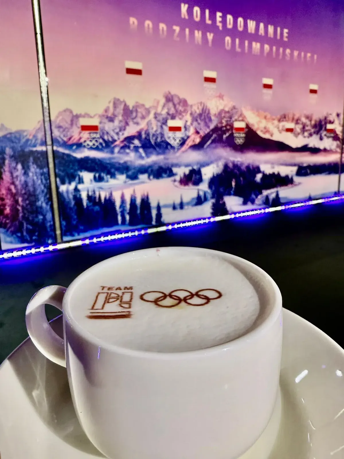 Filiżanka z logo Team PL na piance, w tle grafika z górami