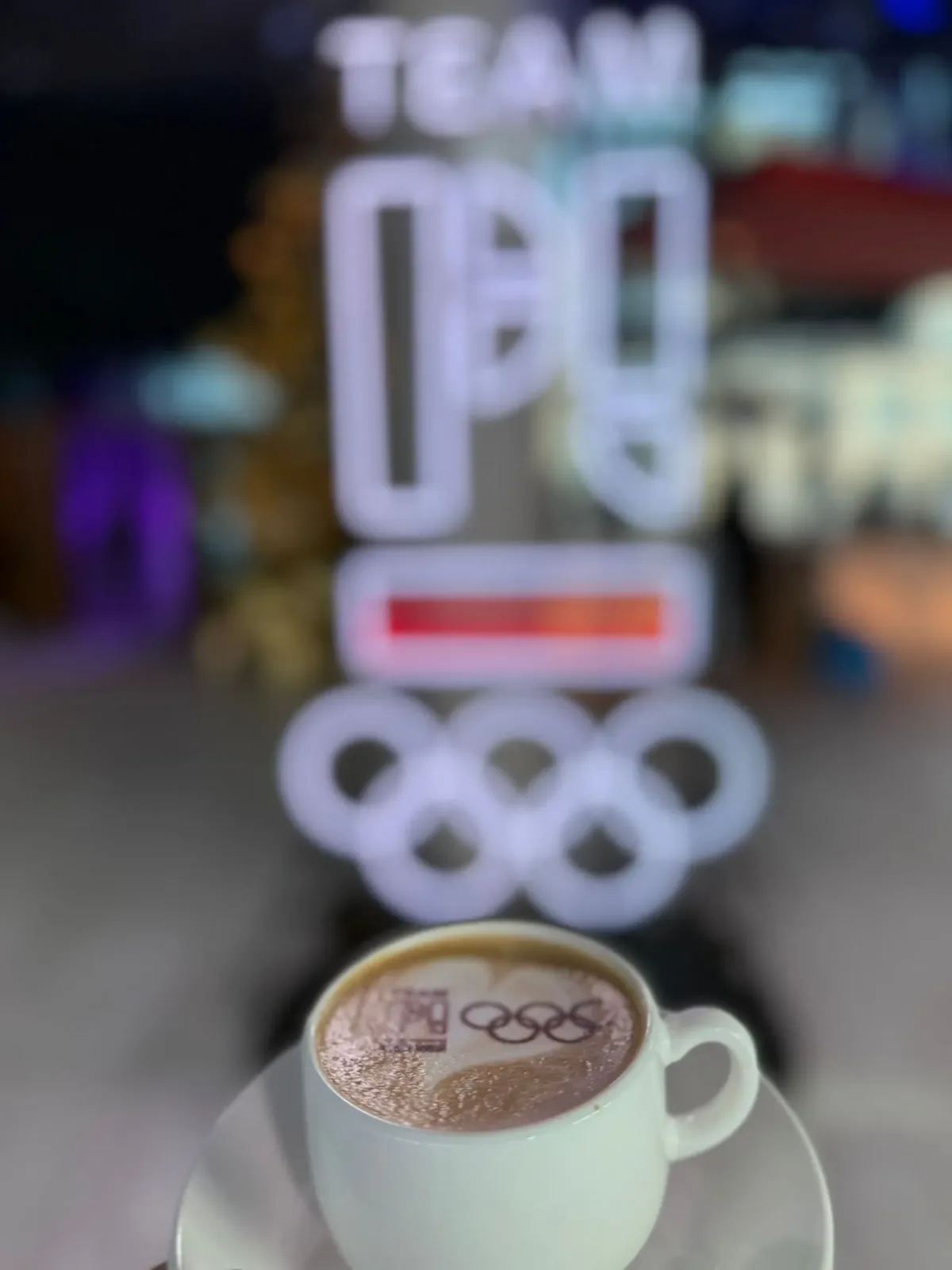 Filiżanka z logo Team PL na piance, w tle świetlne koła olimpijskie