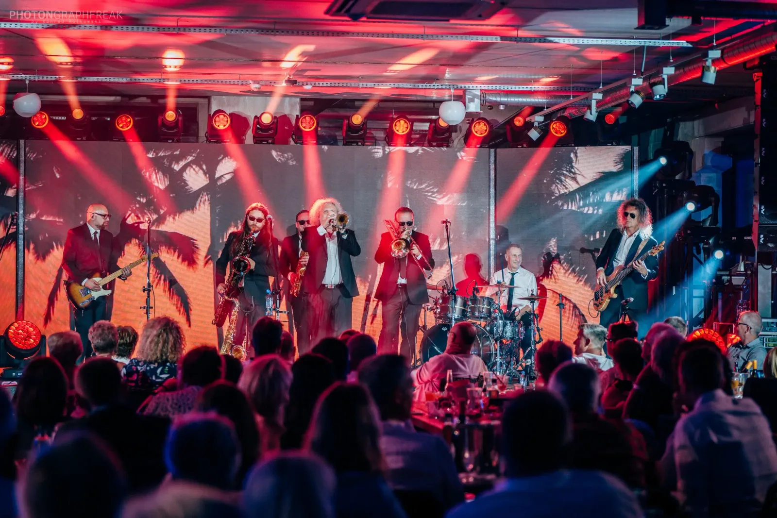 Big band w garniturach gra na scenie w czerwonym świetle, przed nim publiczność przy stołach