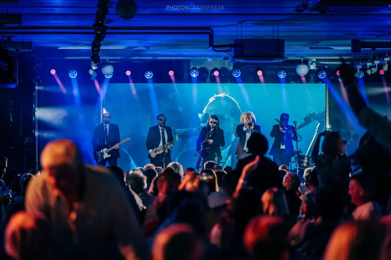 Zespół dęty na scenie w niebieskim świetle, przed sceną tańcząca publiczność