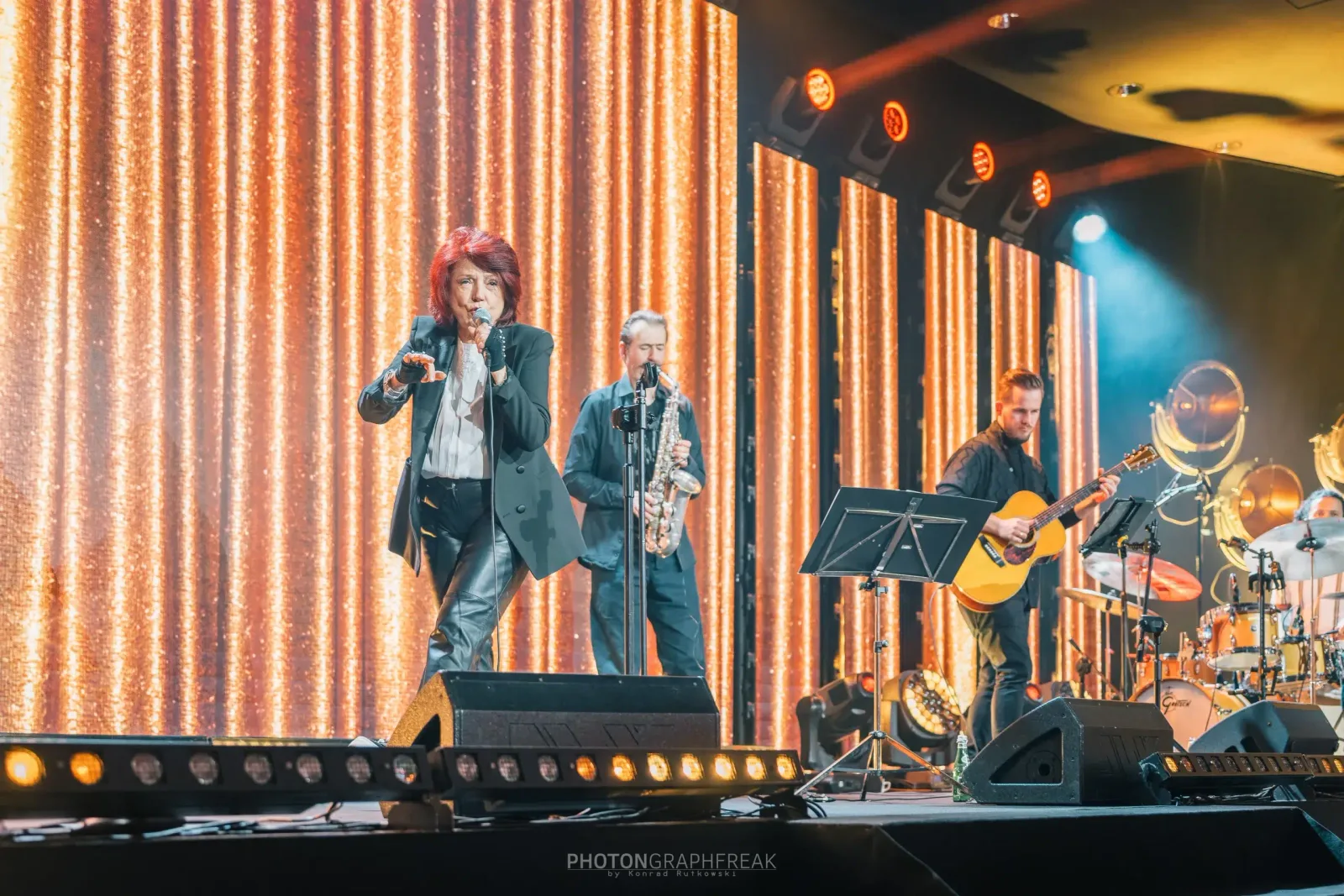 Wokalistka na dużej scenie z saksofonistą i gitarzystą, w tle złota kurtyna świetlna