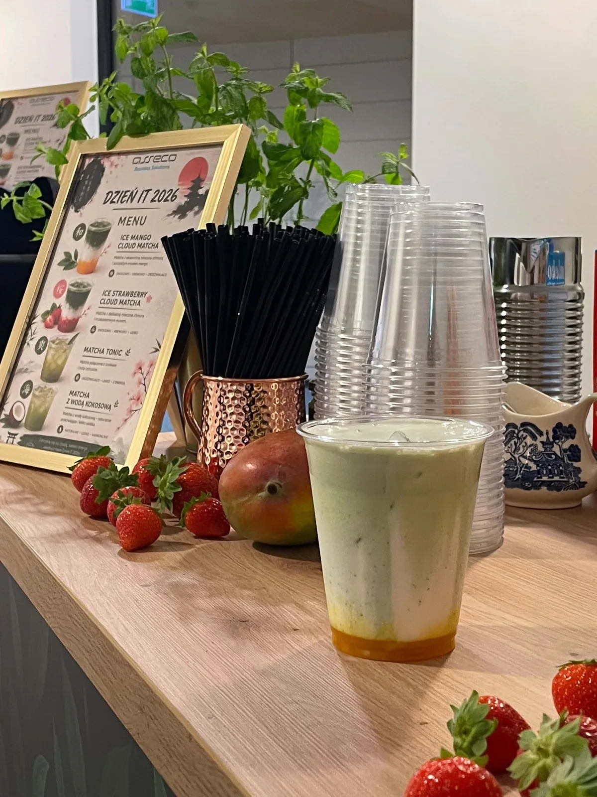 Mrożona matcha z mleczną chmurą i musem mango na barze, obok truskawki, mango i karta menu w ramce