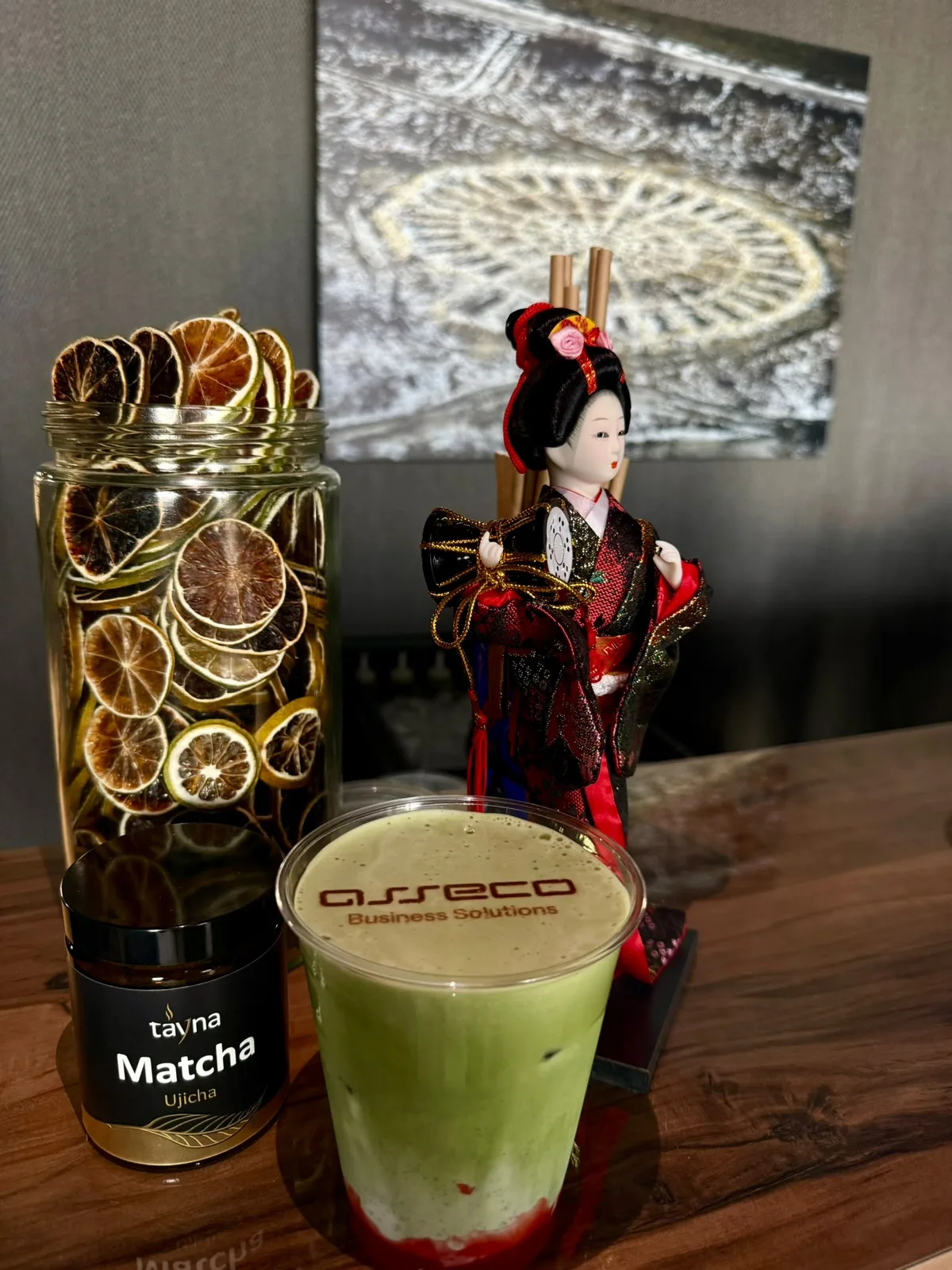Matcha latte z nadrukiem na piance obok słoika matchy, suszonych cytrusów i figurki gejszy