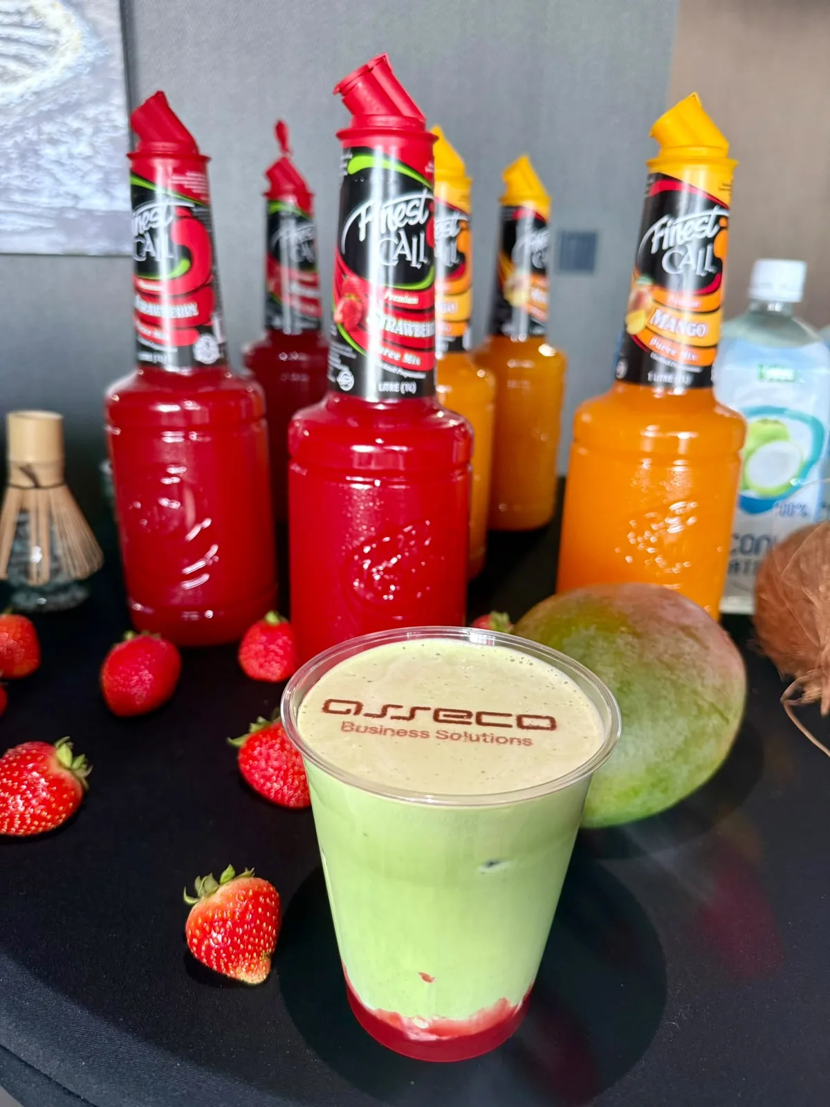 Matcha z musem truskawkowym i logo firmy nadrukowanym na piance, w tle butelki puree z truskawek i mango