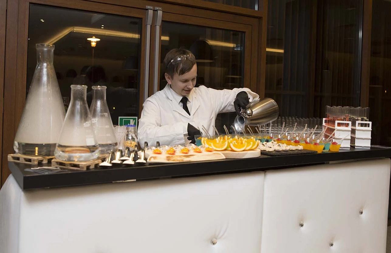 Barman w białym kitlu nalewa drink przy barze molekularnym zastawionym kolbami i plastrami pomarańczy
