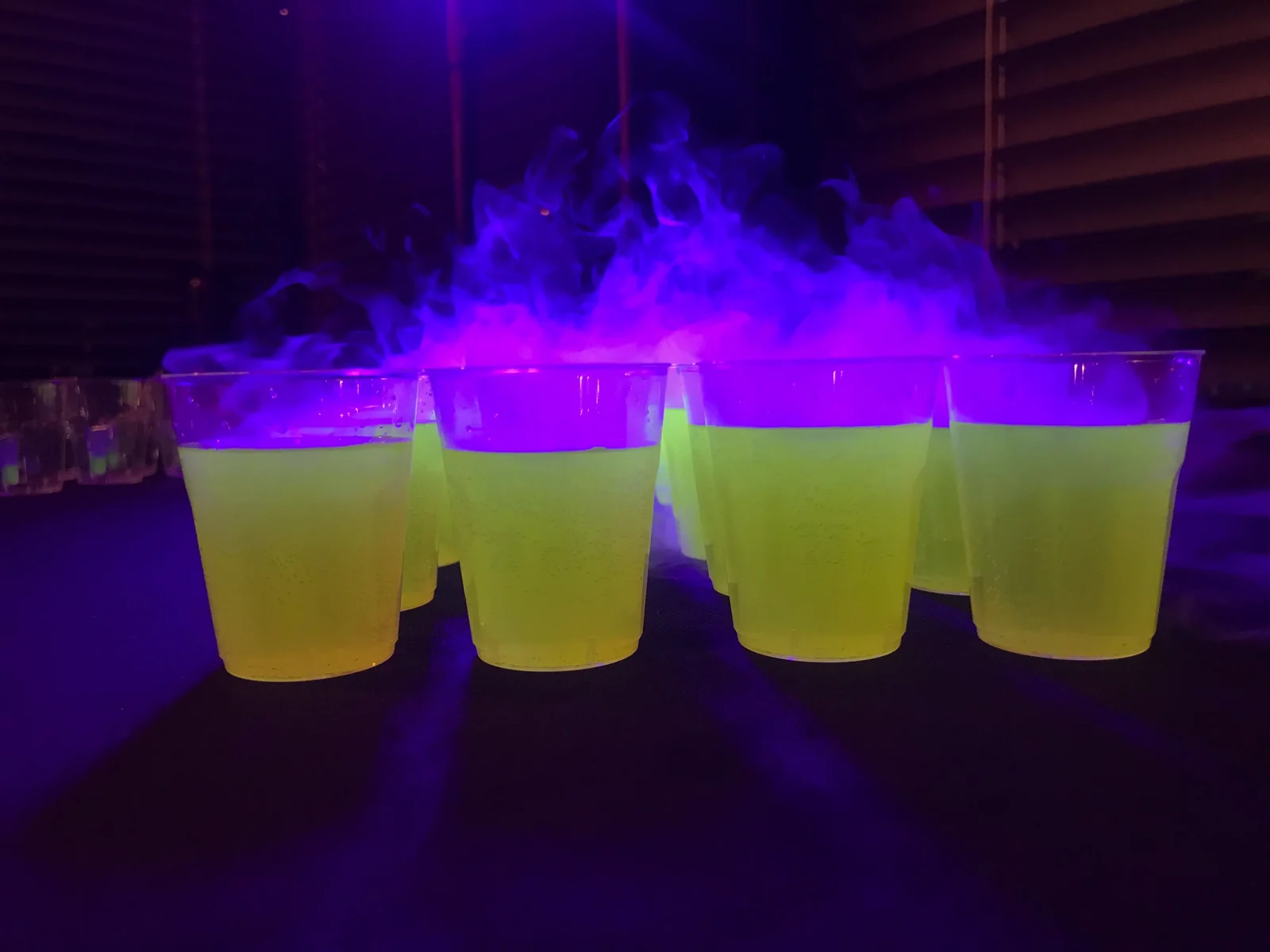 Cztery kubki z żółtozielonym drinkiem w fioletowym świetle UV, spowite dymem