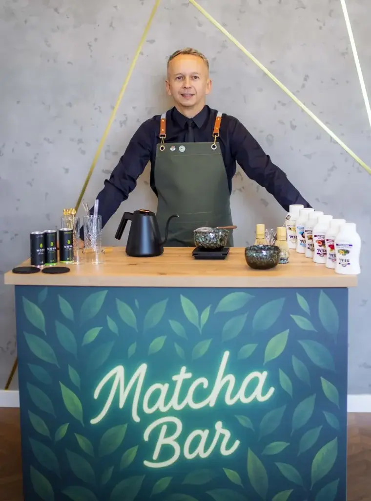 Barista w fartuchu za brandowanym barem z napisem Matcha Bar, na blacie czajnik, misa chawan i syropy